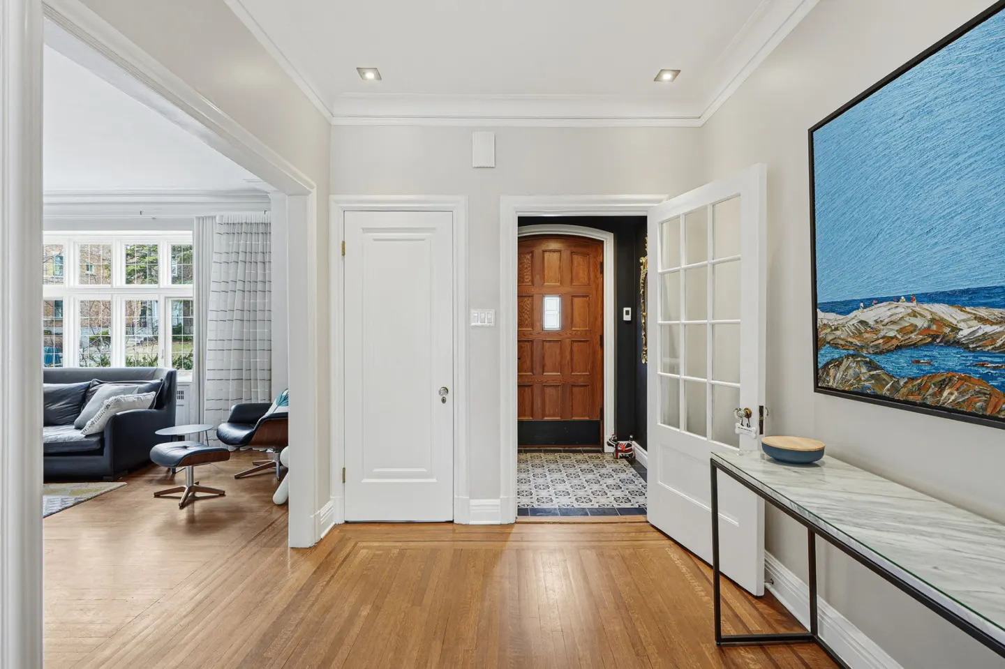 A bright foyer with hardwood floors, white trim, and a marble-topped console table. A living room is visible through an open doorway.