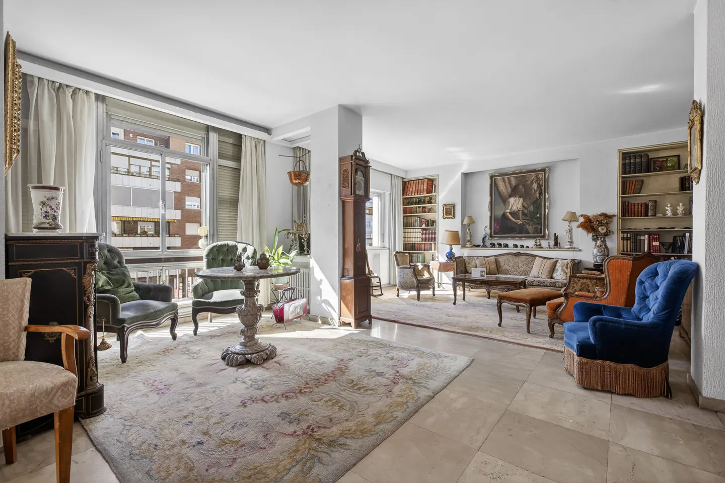 Bright living room with marble floors, floral rug, and antique furniture. Large windows with white curtains let in natural light.