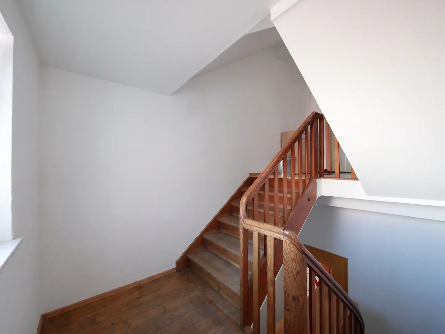 Interior view of a wooden staircase with white walls and ceiling. The stairs have a wooden railing and lead to an upper level.