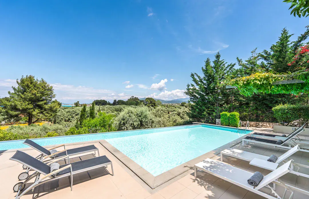A rectangular swimming pool with lounge chairs on a tiled patio, surrounded by lush trees under a bright blue sky.