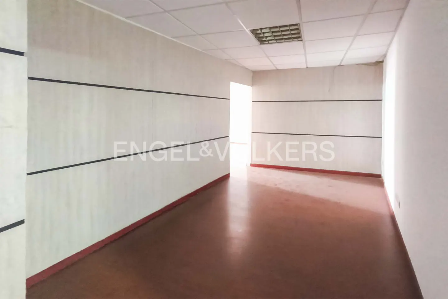 Empty office space with white walls, brown floor, and black horizontal lines. Engel & Volkers logo is visible on the wall.