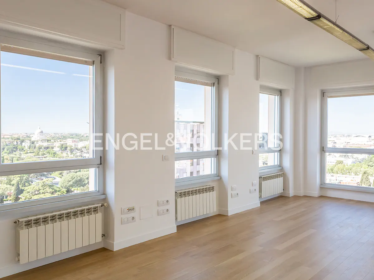 Bright, empty room with light wood floors, white walls, and four large windows with city views. Radiators under each window.