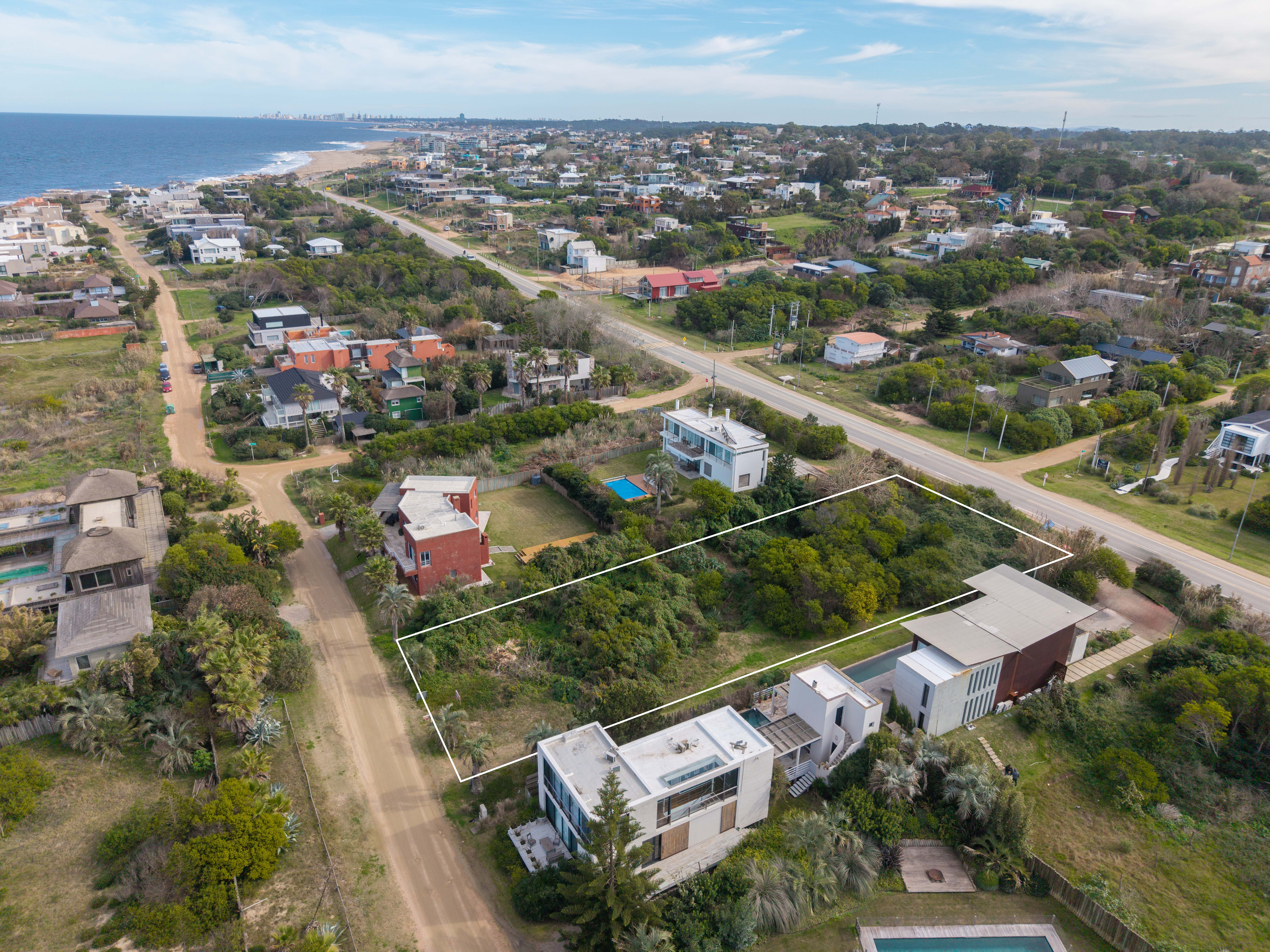 Aerial view of a coastal neighborhood with a highlighted vacant lot surrounded by modern homes and lush greenery near the sea. [AI-generated text]
