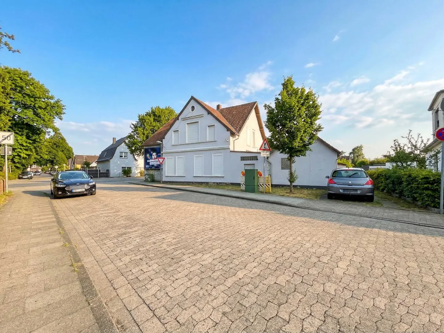 Exterior view of a two-story white house with a brown roof on a paved street. A black car is driving on the street, and a gray car is parked in front of the house.