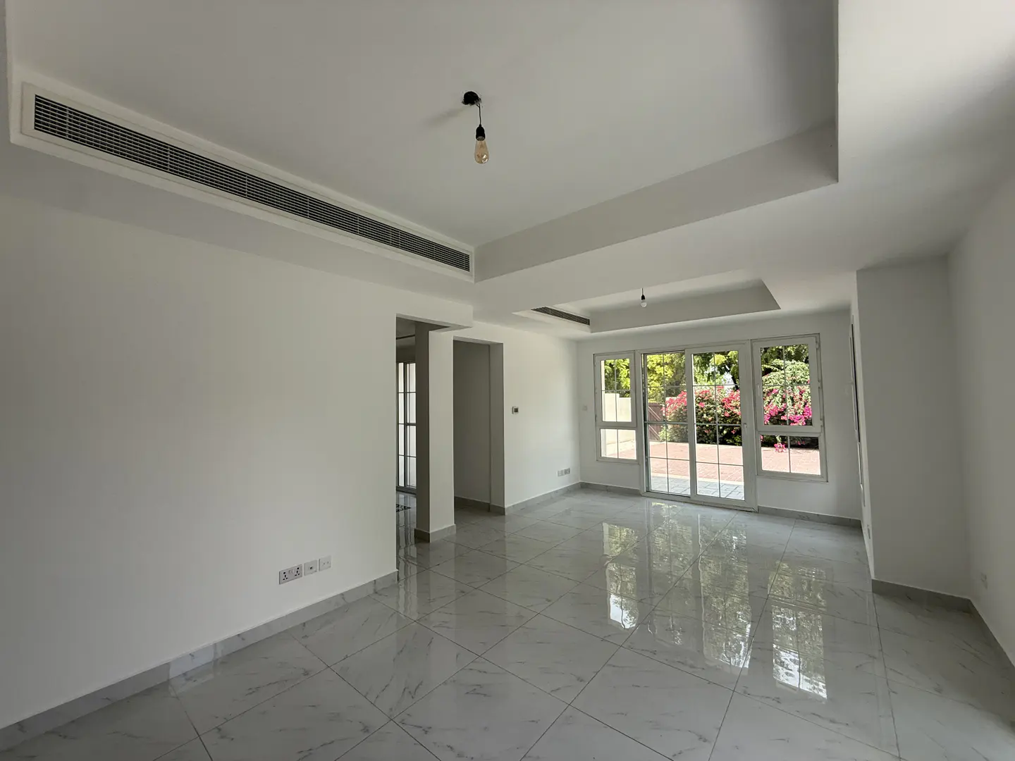 Bright, empty room with white walls and marble floors. Sliding glass doors lead to a garden with pink flowers.