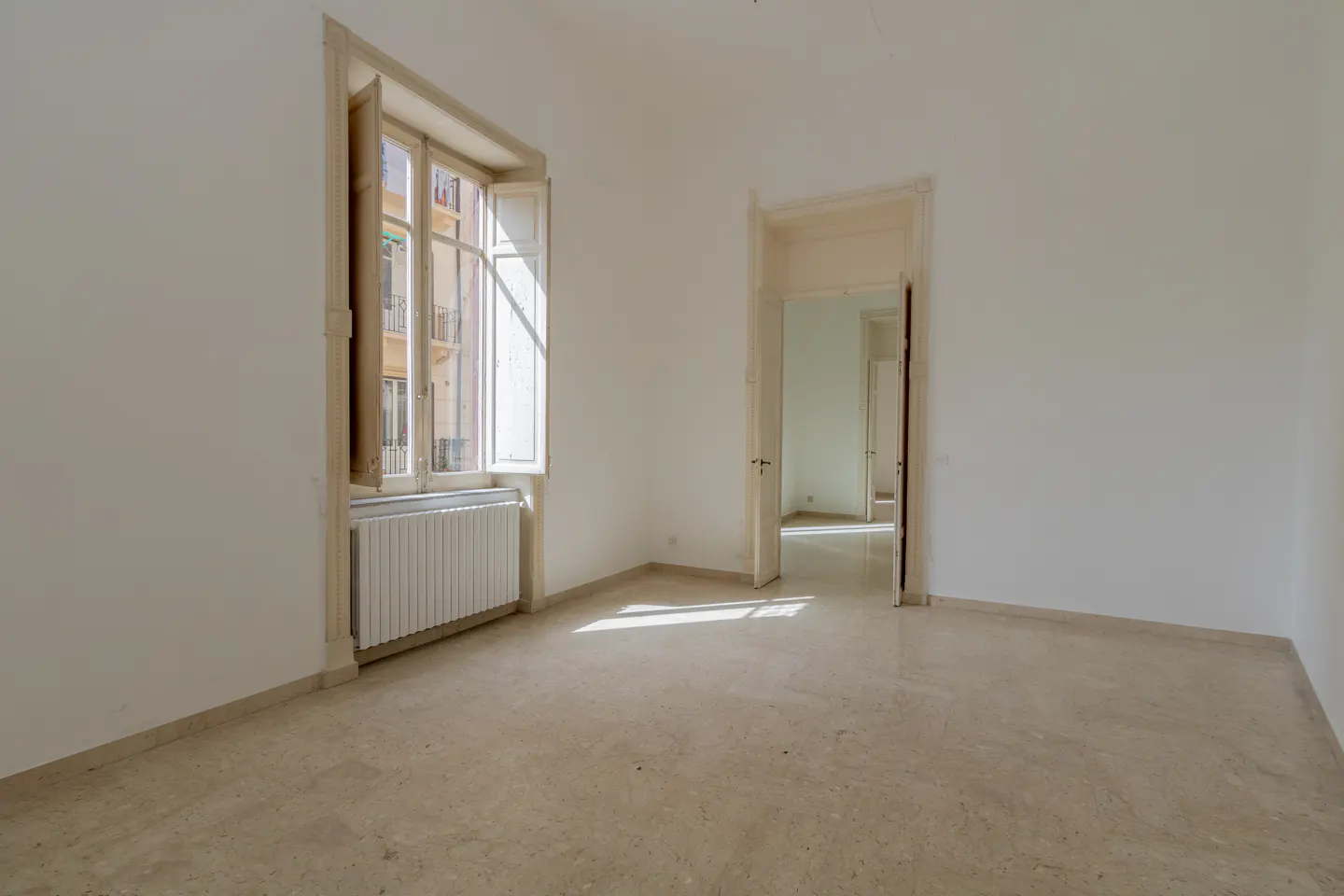 An empty, bright room with a window and an open doorway. The walls are white, and the floor is beige marble. Sunlight streams through the window.