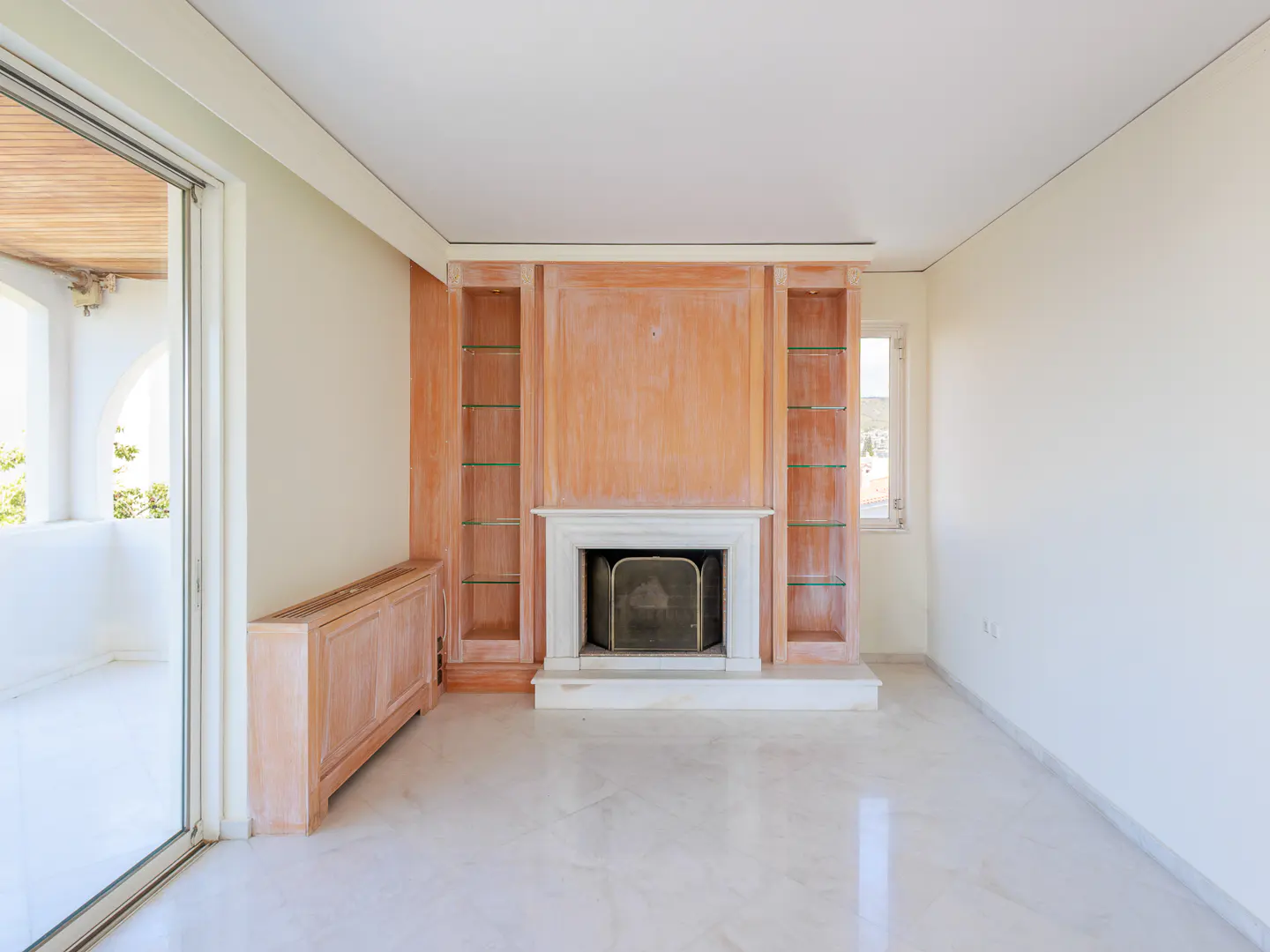 Bright, empty room with marble floors, a fireplace with a black screen, and built-in wooden shelves. A sliding glass door leads to a balcony.