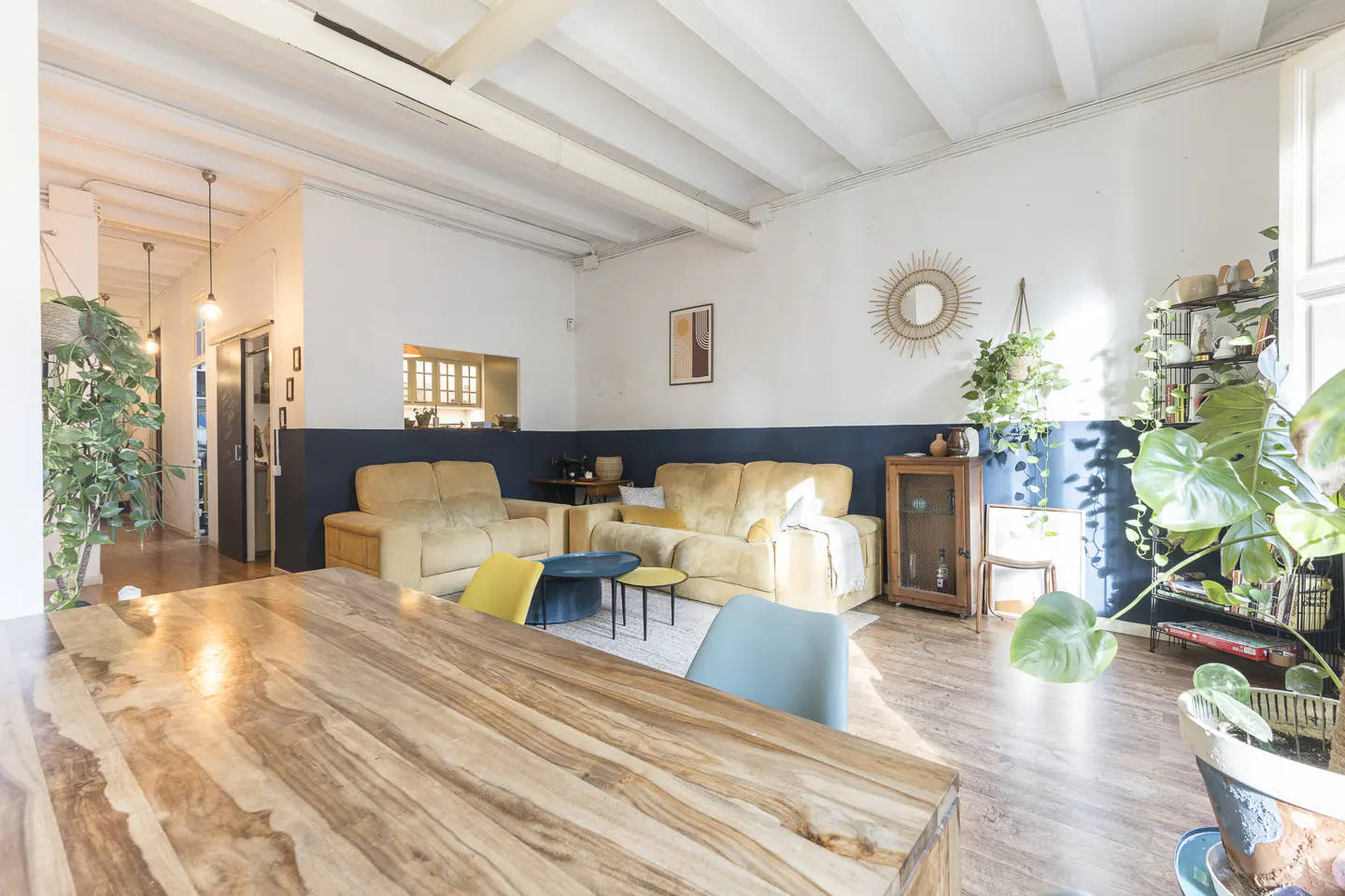 Bright, open living room with wood floors, white walls, and blue accent. Tan sofas, wood table, and potted plants add warmth.