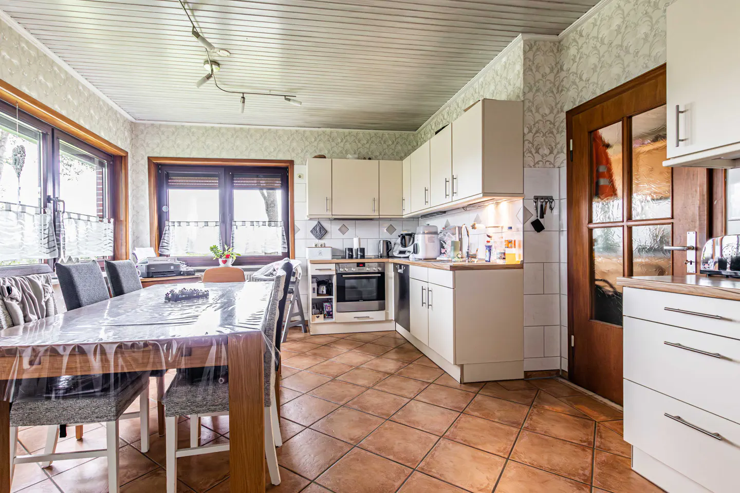 A kitchen with a dining area features white cabinets, brown tile floors, and a wooden table with chairs. Natural light streams through the windows.