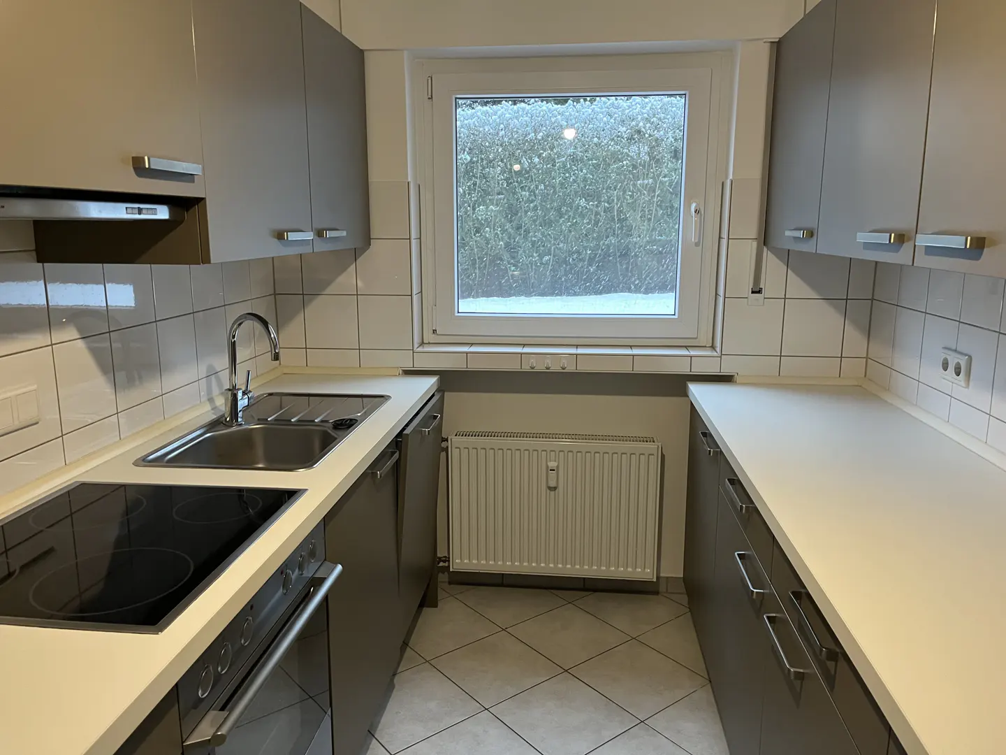 A galley kitchen with gray cabinets, white countertops, and a stainless steel sink. A window overlooks a snowy outdoor scene.