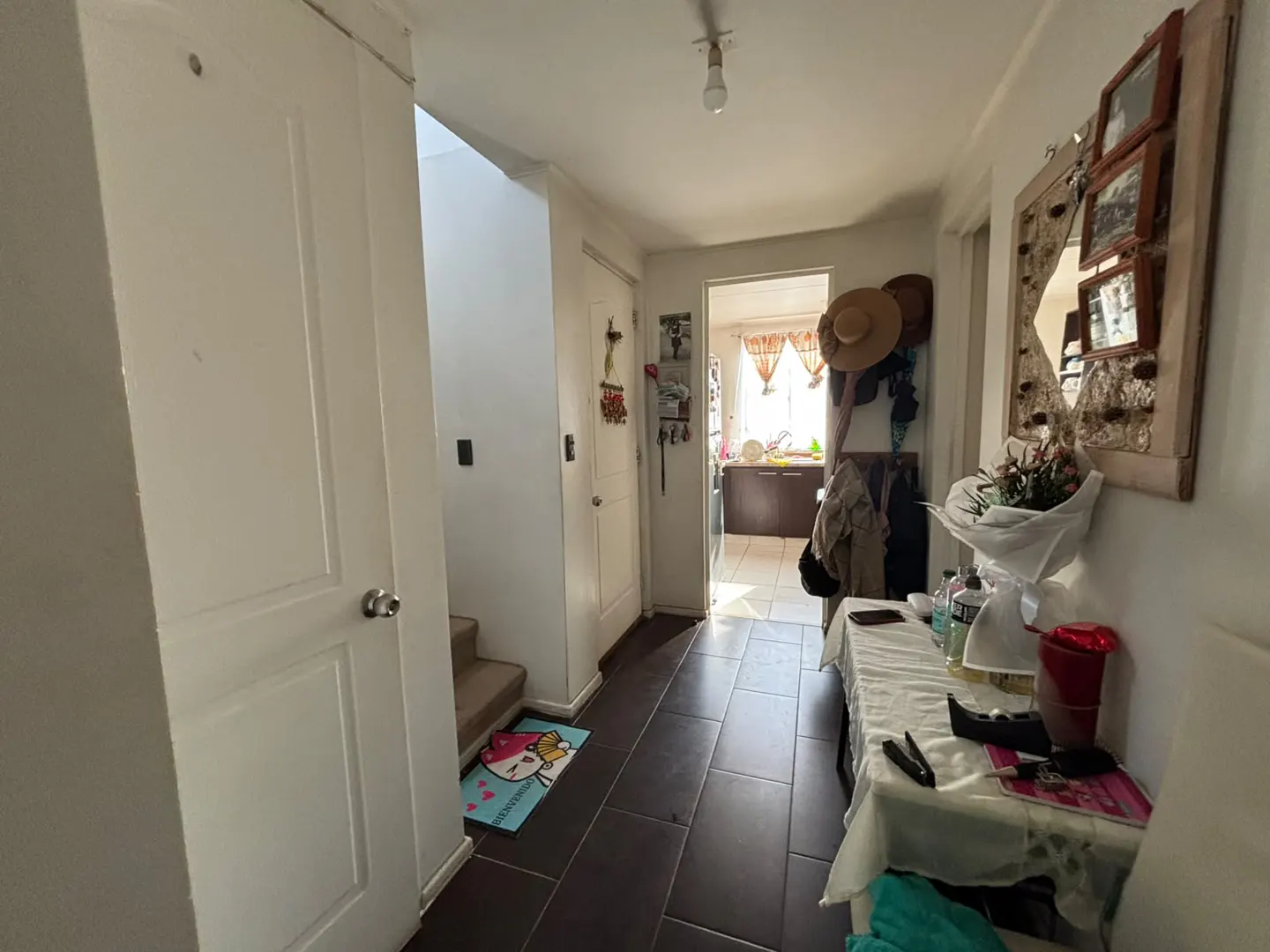 A hallway with dark tile floors, a white table with a bouquet, a wall mirror, and a doorway leading to a bright kitchen.