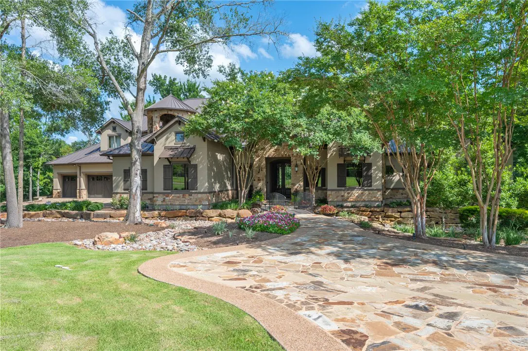 A tan stone house with dark shutters and a stone driveway is surrounded by green trees and grass.