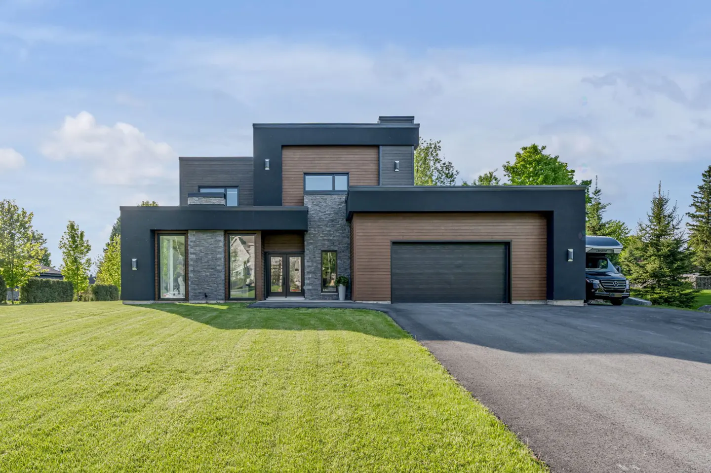 Modern two-story home with black and wood siding, stone accents, a large garage, and a green lawn under a blue sky.