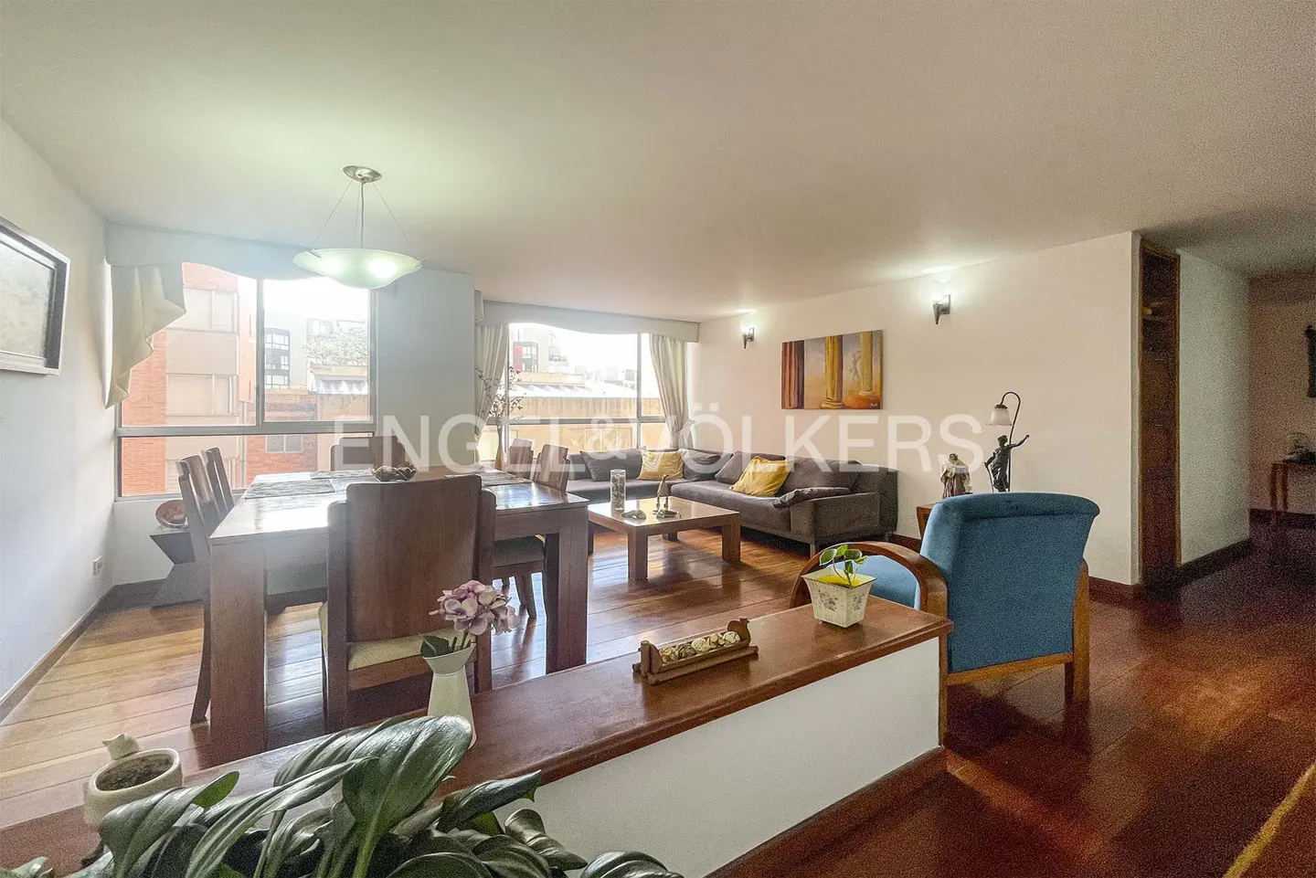 Open-concept living and dining area with hardwood floors, a gray sofa, and a wooden dining table. Large windows provide natural light.