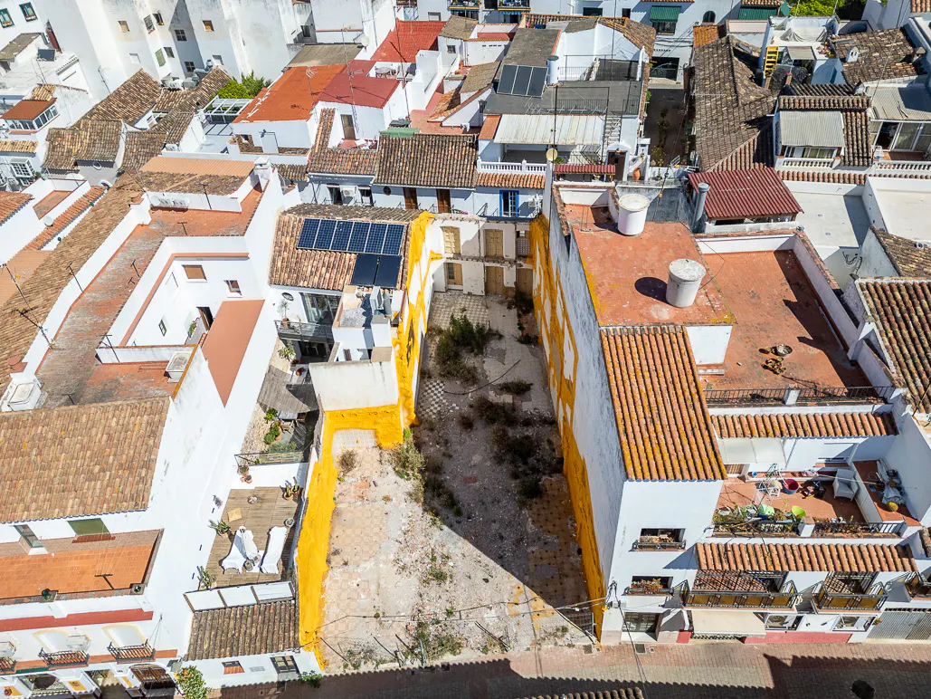 Aerial view of a vacant lot surrounded by white buildings with orange trim and terracotta roofs in a sunny, urban setting.