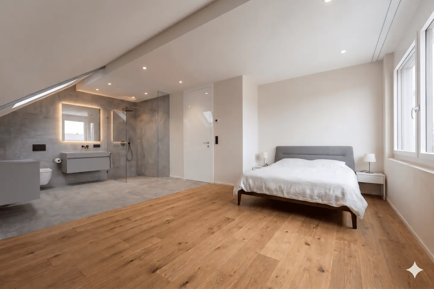 Open-concept bedroom and bathroom with wood floors, white walls, and a gray tiled bathroom with a glass shower. A bed is on the right.