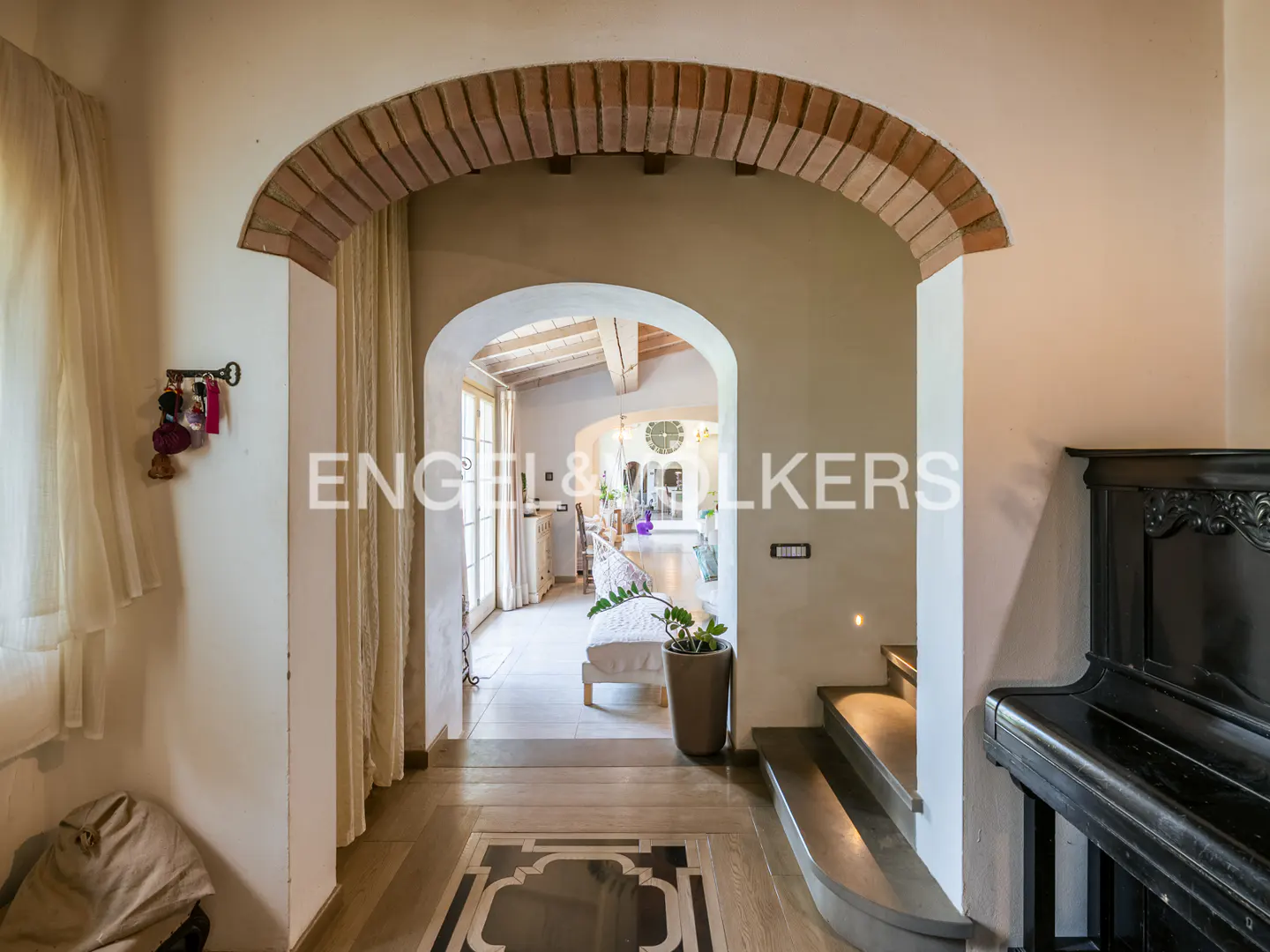 Interior view of a home with brick archways leading to a bright living space, a black piano on the right, and neutral tones throughout.