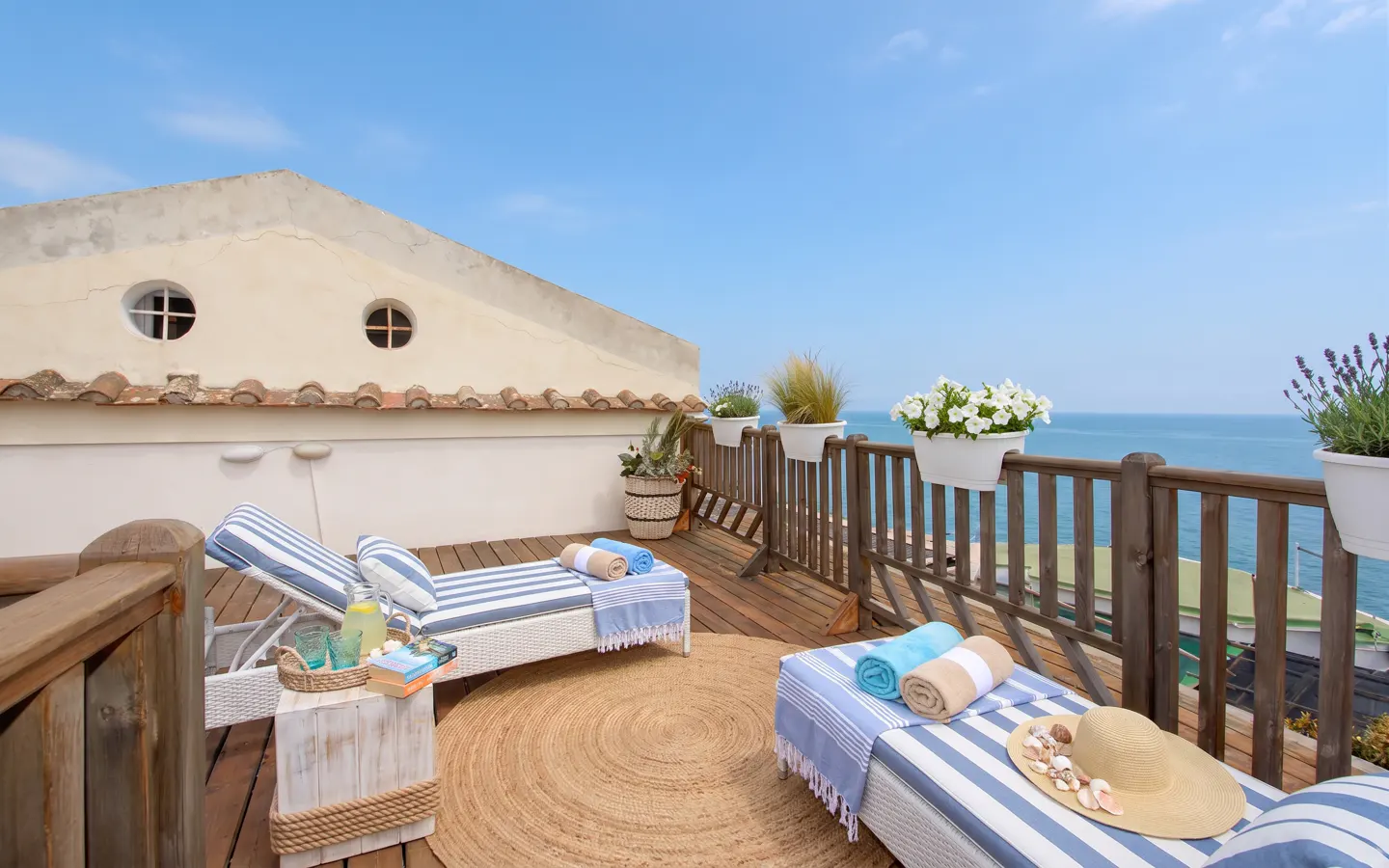 Rooftop patio with ocean view. Two striped lounge chairs, a round rug, and a wooden railing with flower boxes.