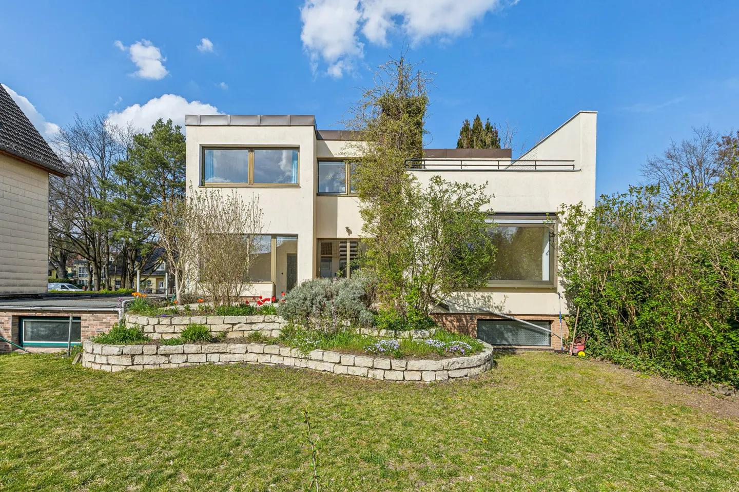 Modern two-story house with large windows, stone retaining wall, and green lawn under a blue sky.