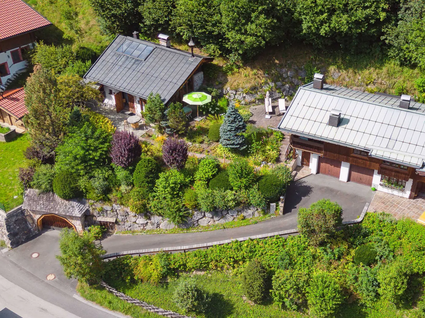 Aerial view of two houses with gray roofs, surrounded by lush green trees and bushes. A stone wall and driveway lead to the houses.