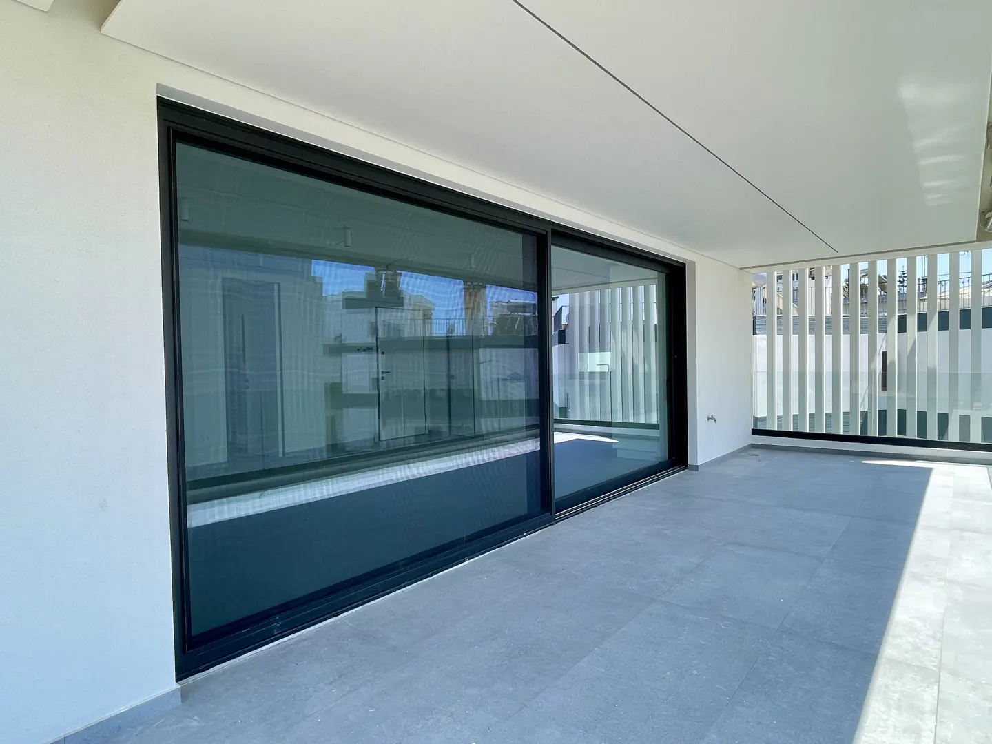 A modern balcony with gray tile flooring, a white wall, and a large sliding glass door with a black frame.