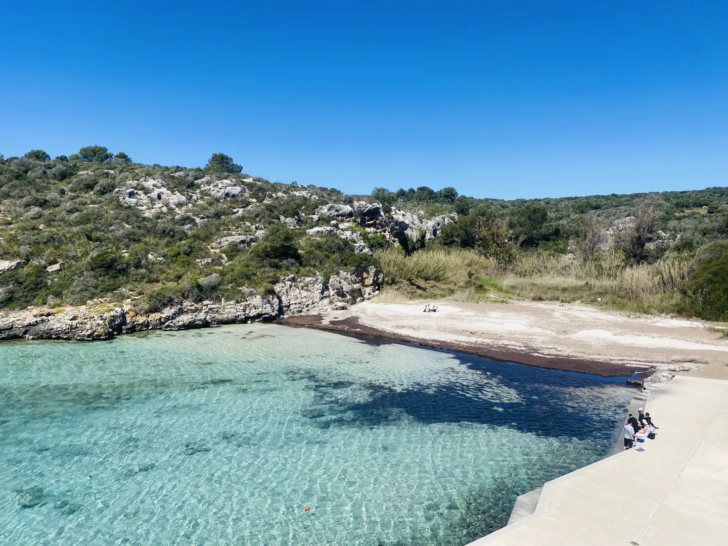 Scenic view of a cove with clear turquoise water, a sandy beach, and rocky hills under a bright blue sky. People are on the pier.