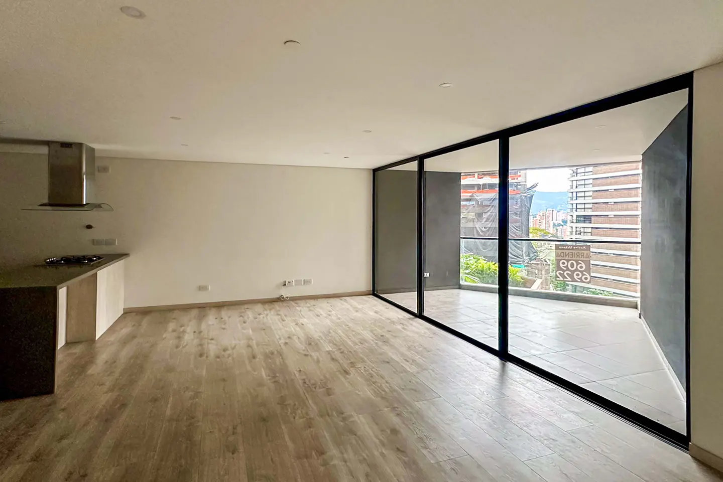 An empty, bright apartment with wood floors and a kitchen island. Large black-framed windows offer a city view.