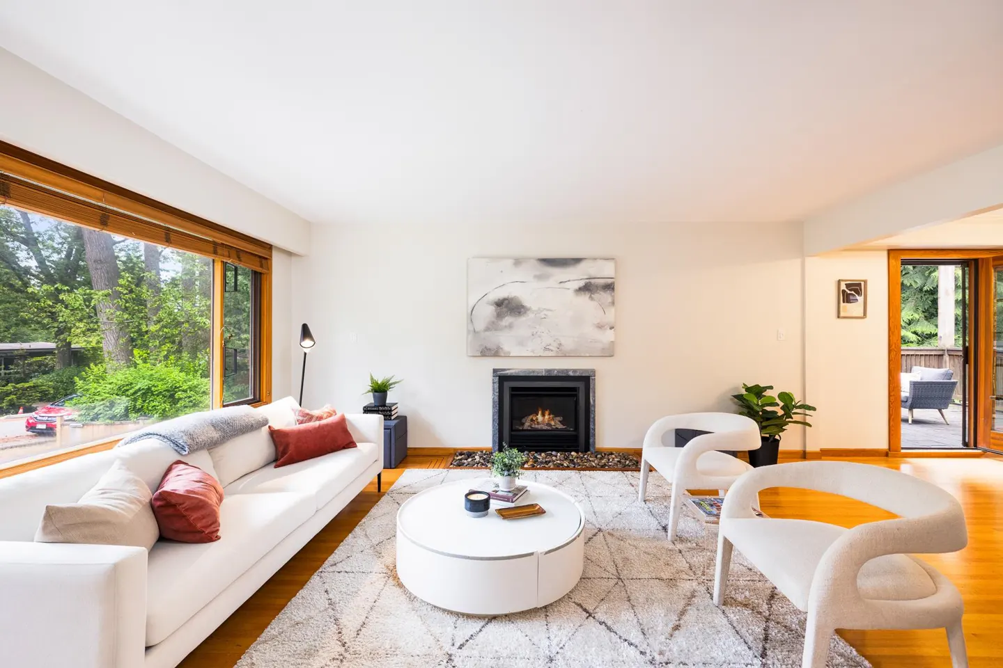 Bright living room with white sofa, two modern chairs, round coffee table, and fireplace, overlooking a wooded backyard.
