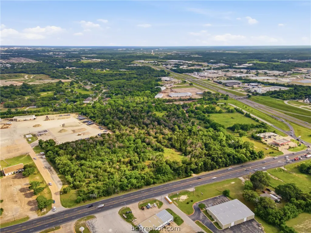 Aerial view of a wooded lot next to a highway, with buildings and trees in the background under a blue sky.