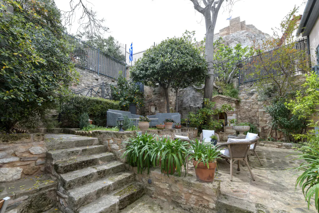 Outdoor patio with stone steps, potted plants, and seating areas. A Greek flag is visible in the background, near a stone building on a hill.