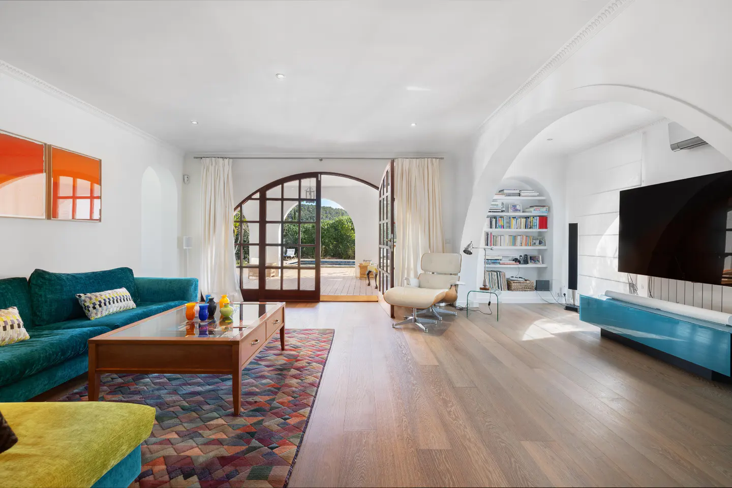 Bright living room with teal sofa, colorful patterned rug, wooden coffee table, and arched doors opening to a patio.