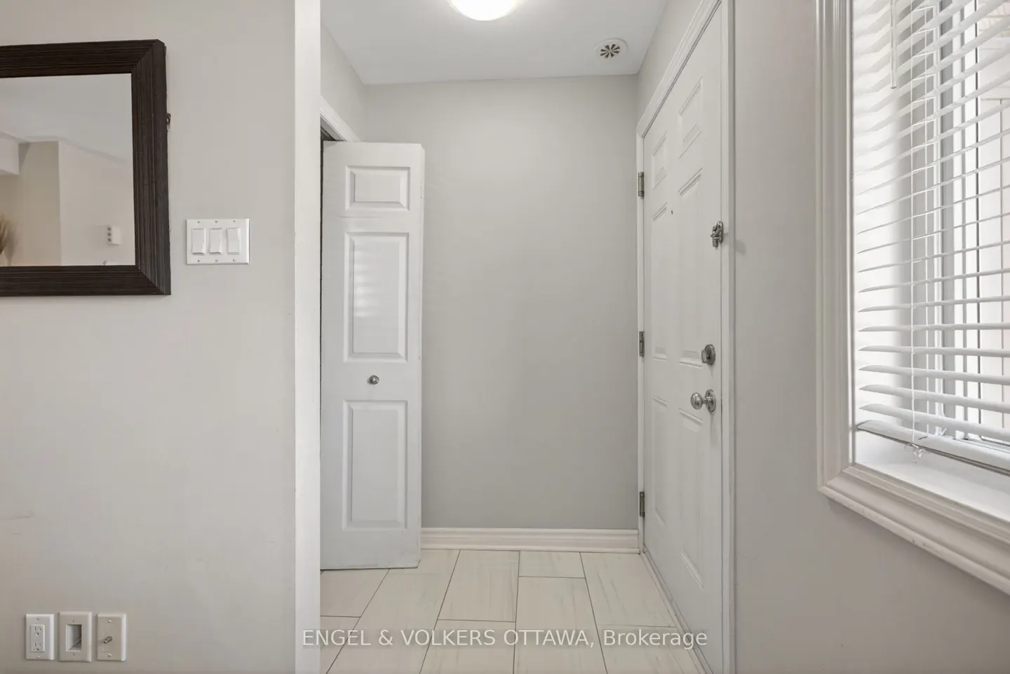 Entryway with white walls, tile floor, and doors. A dark-framed mirror hangs on the left wall. A window with blinds is on the right.
