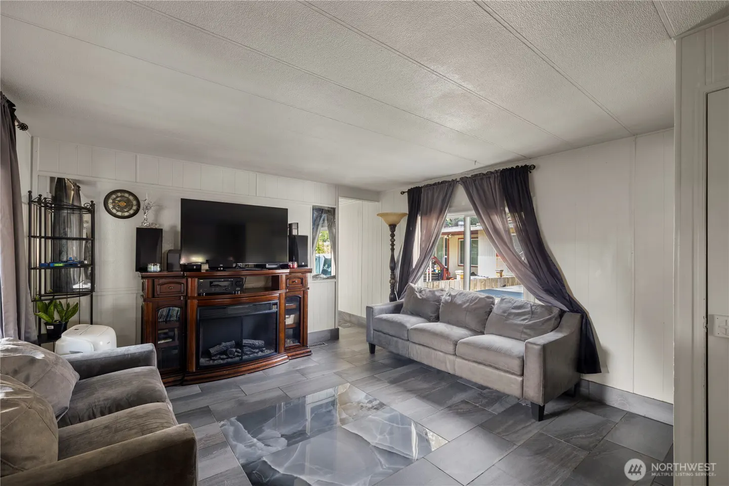 Living room with gray sofas, a fireplace with a TV above, and gray tile flooring. Curtains frame a window with an outside view.