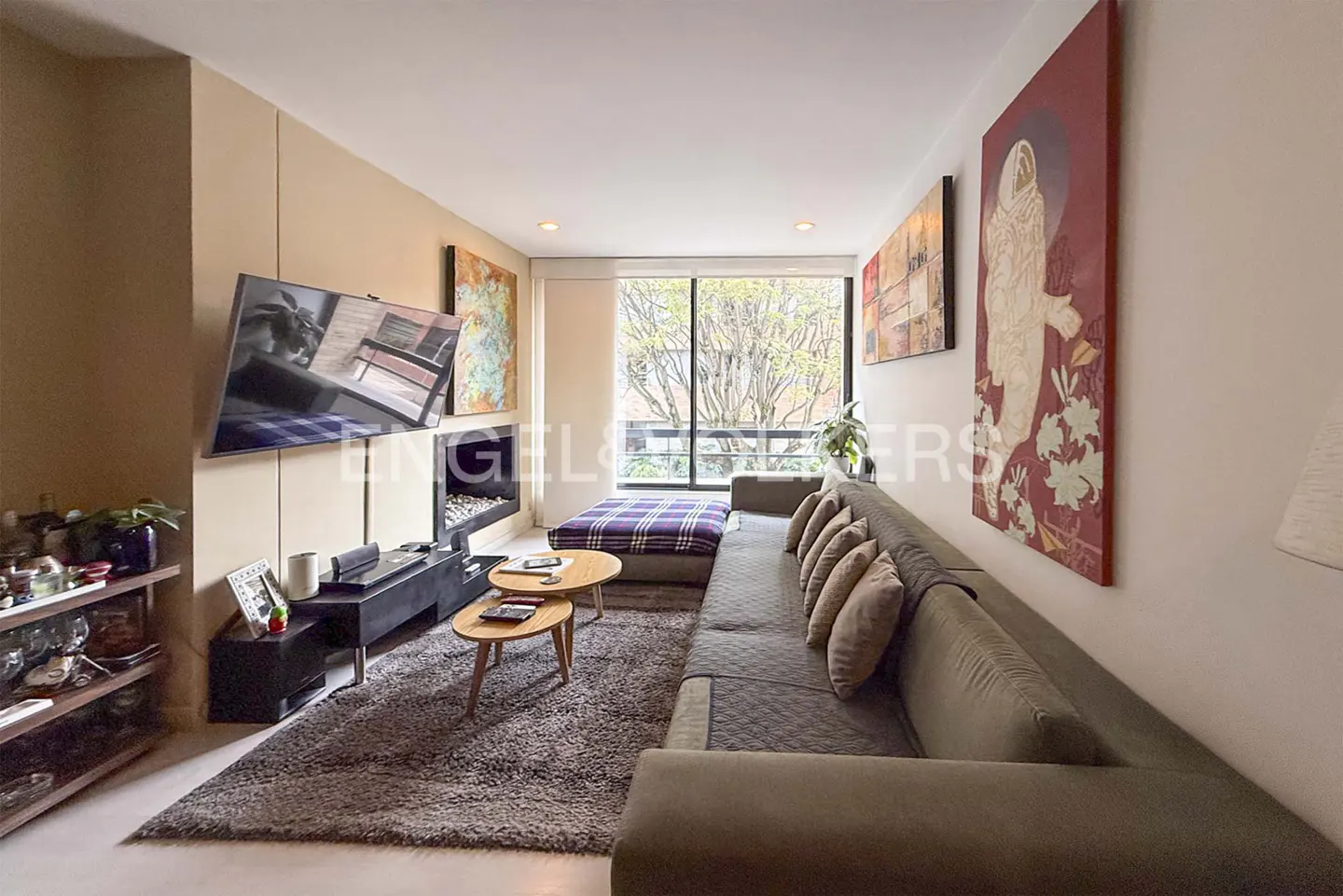 Living room with a gray sectional sofa, a TV, a fireplace, and a large window with a view of trees.