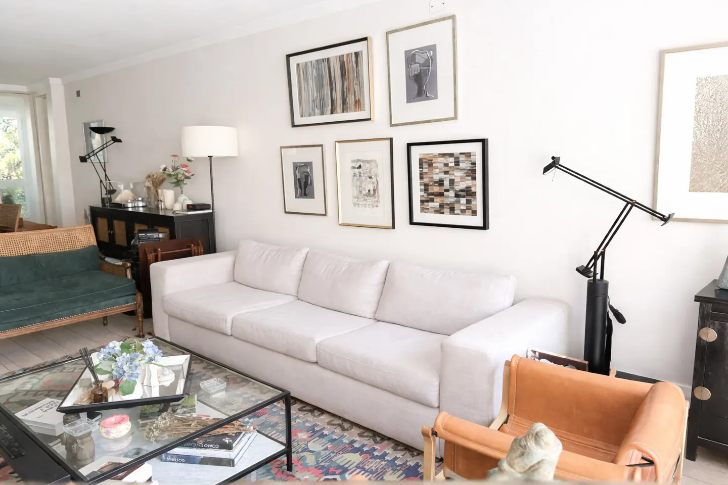 Bright living room with a white sofa, glass coffee table, and gallery wall. A green settee and tan chair add color.