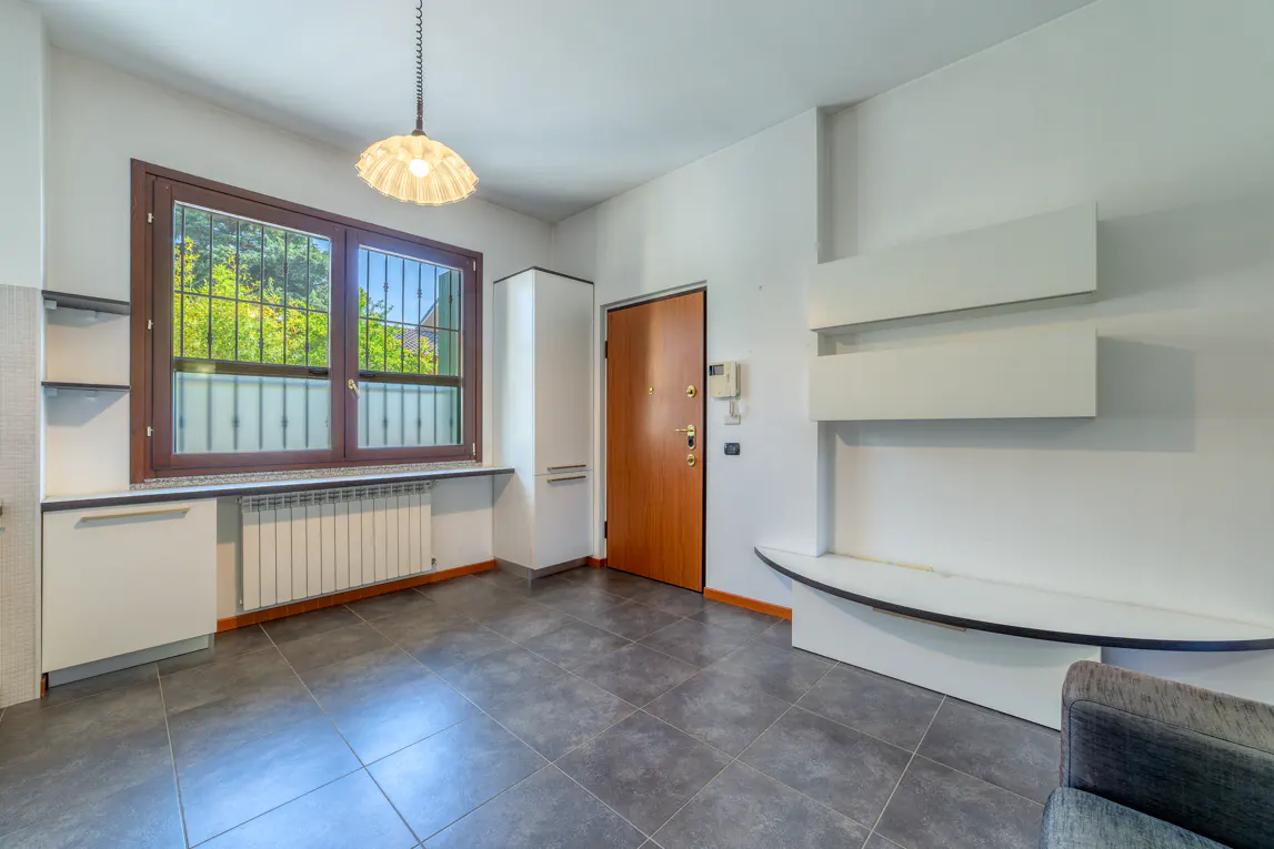 A bright room with gray tile floors, white walls, a brown door, and a window with a view of green trees.