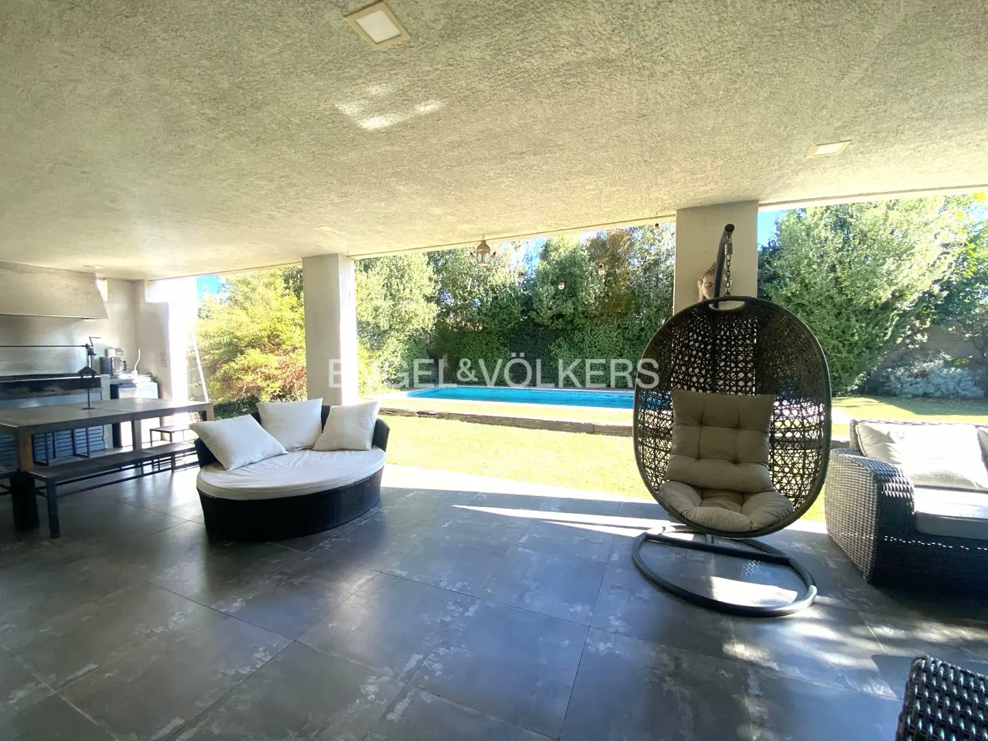 Covered patio with a round daybed, hanging egg chair, and dining table overlooking a backyard swimming pool.
