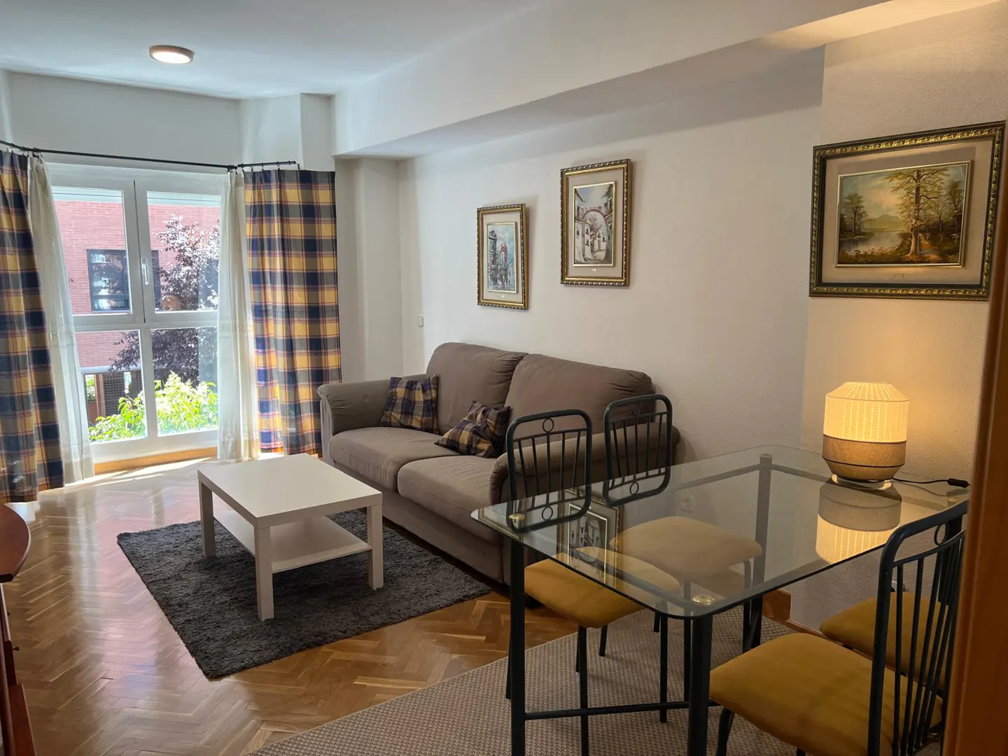 Living room with a brown sofa, white coffee table on a gray rug, and a glass dining table with yellow chairs. Plaid curtains frame a bright window.