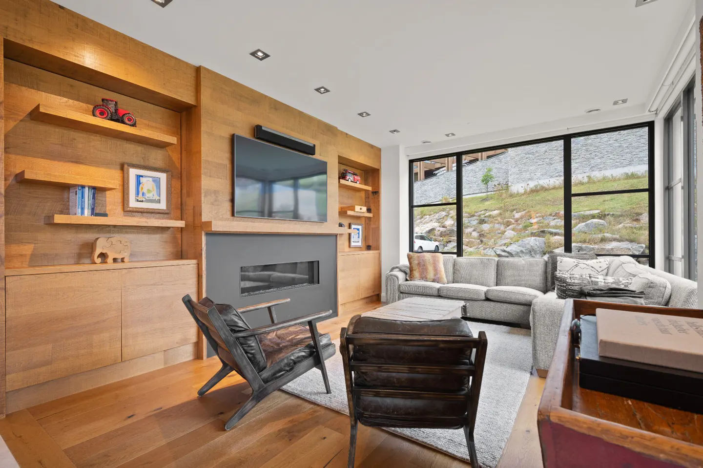 Living room with wood paneled wall, TV, fireplace, gray sofa, and black framed windows with a view of a rocky hillside.