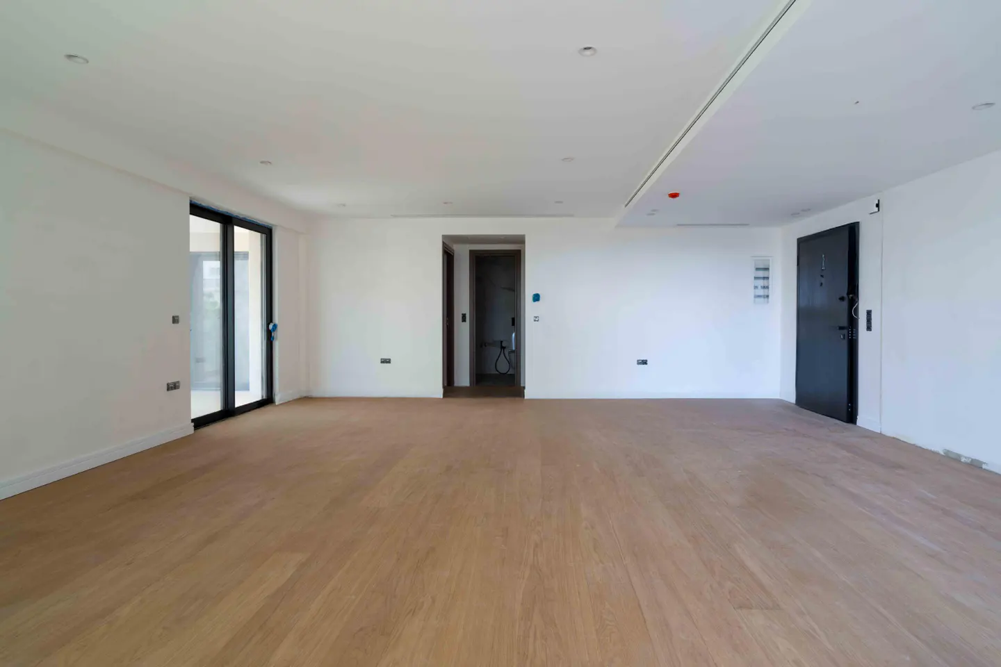 Bright, empty room with light wood floors, white walls, and a black door. A bathroom is visible through a doorway in the back wall.
