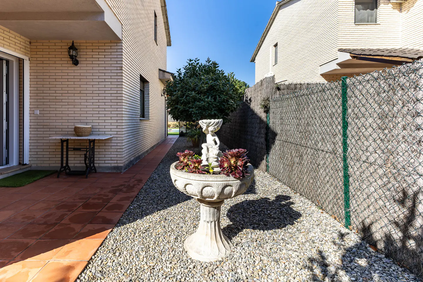 Outdoor view of a garden with a stone fountain, plants, and gravel between two houses under a blue sky.