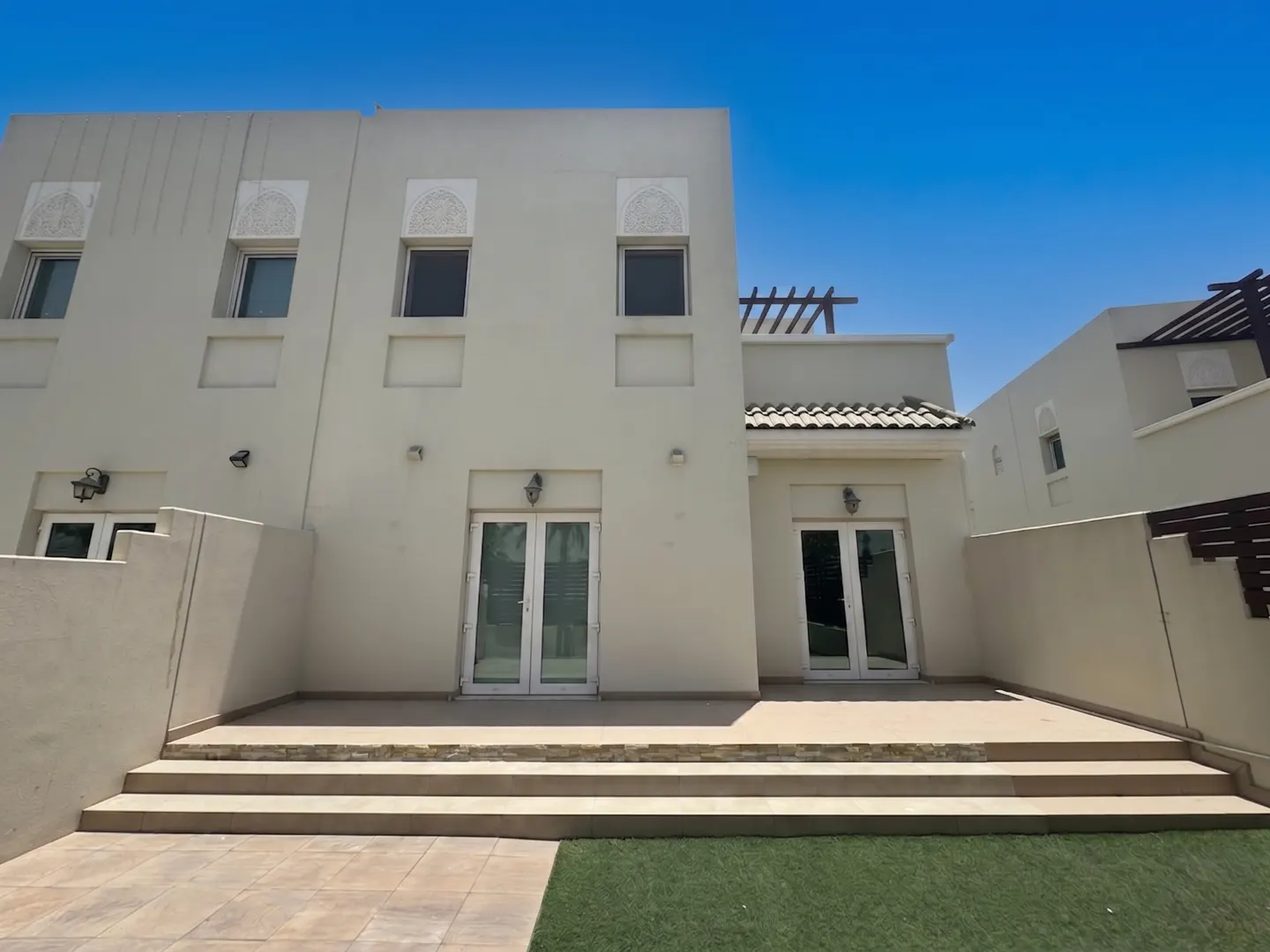 Backyard view of a two-story beige house with white trim, double doors, and a stone patio with grass. Blue sky above.