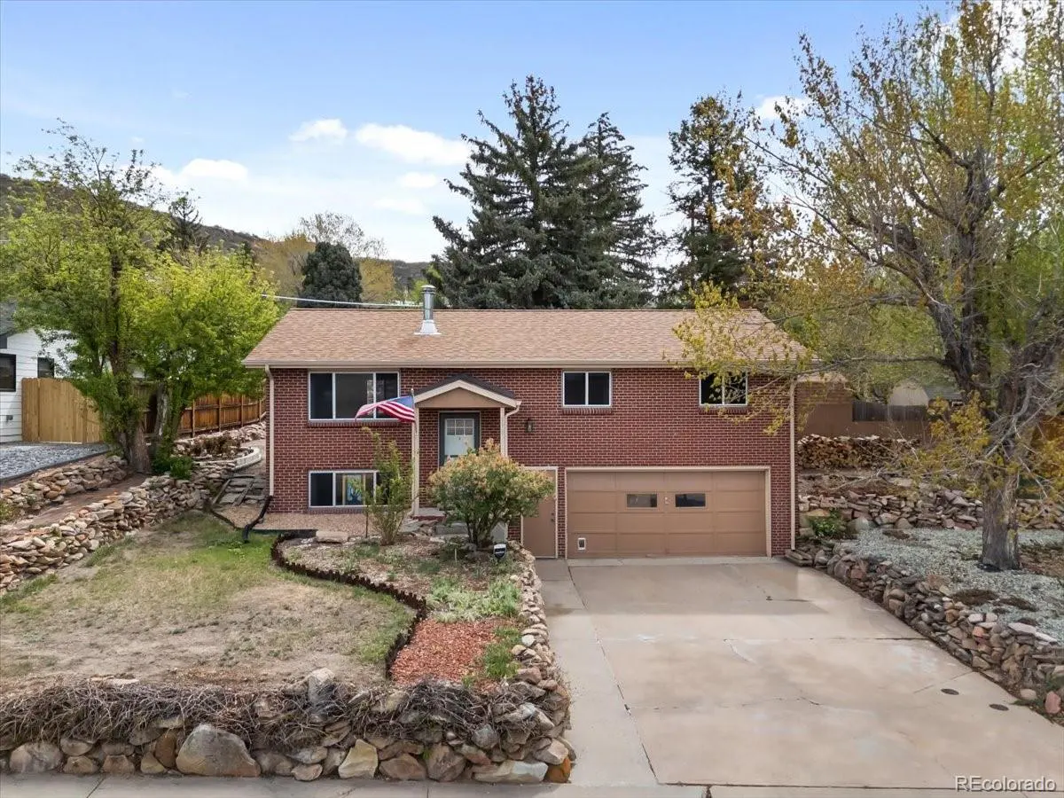 Two-story brick house with brown roof and garage. American flag hangs near the front door. Stone retaining walls line the yard.