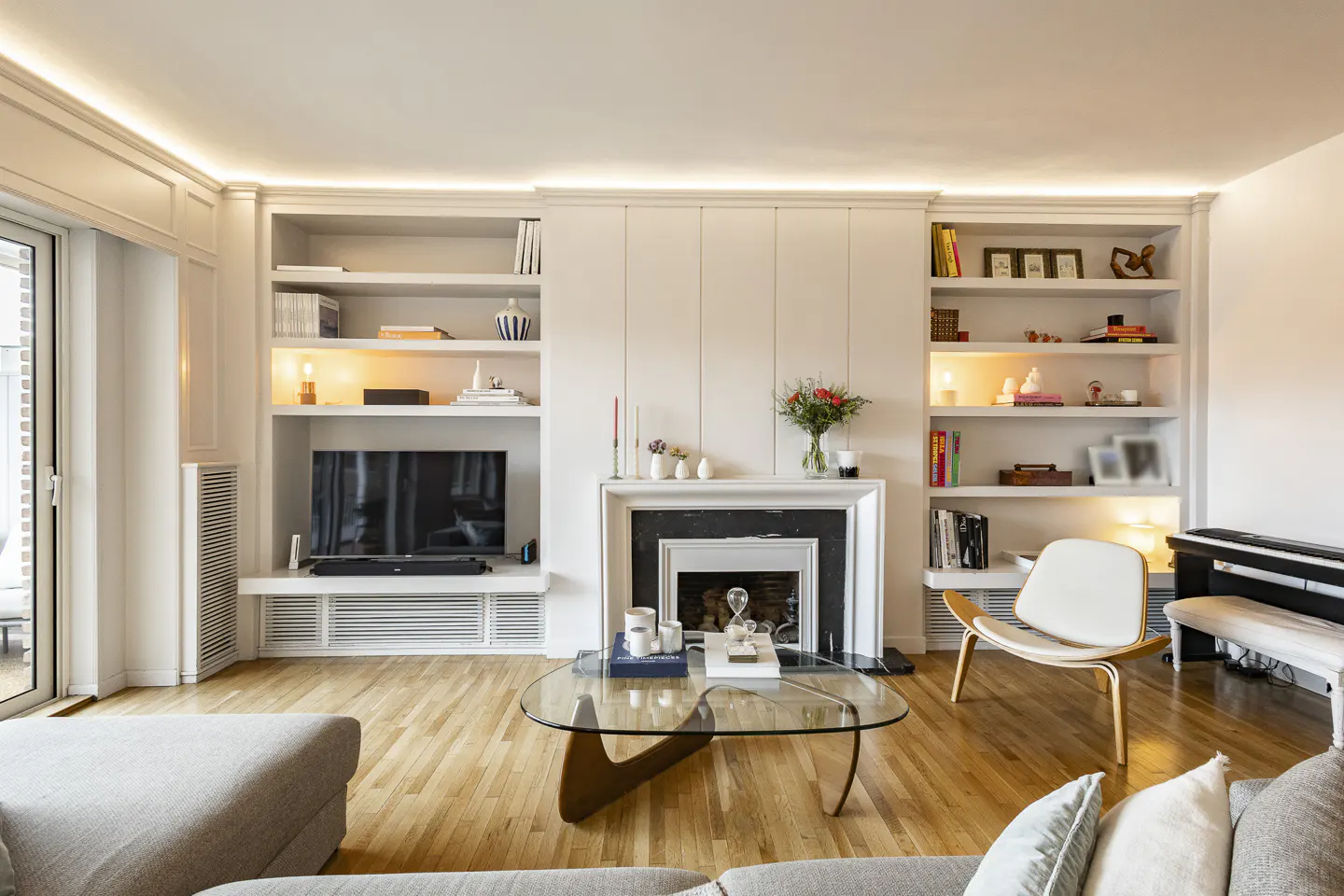 Bright living room with hardwood floors, a glass coffee table, and a white built-in bookcase with a TV and fireplace.