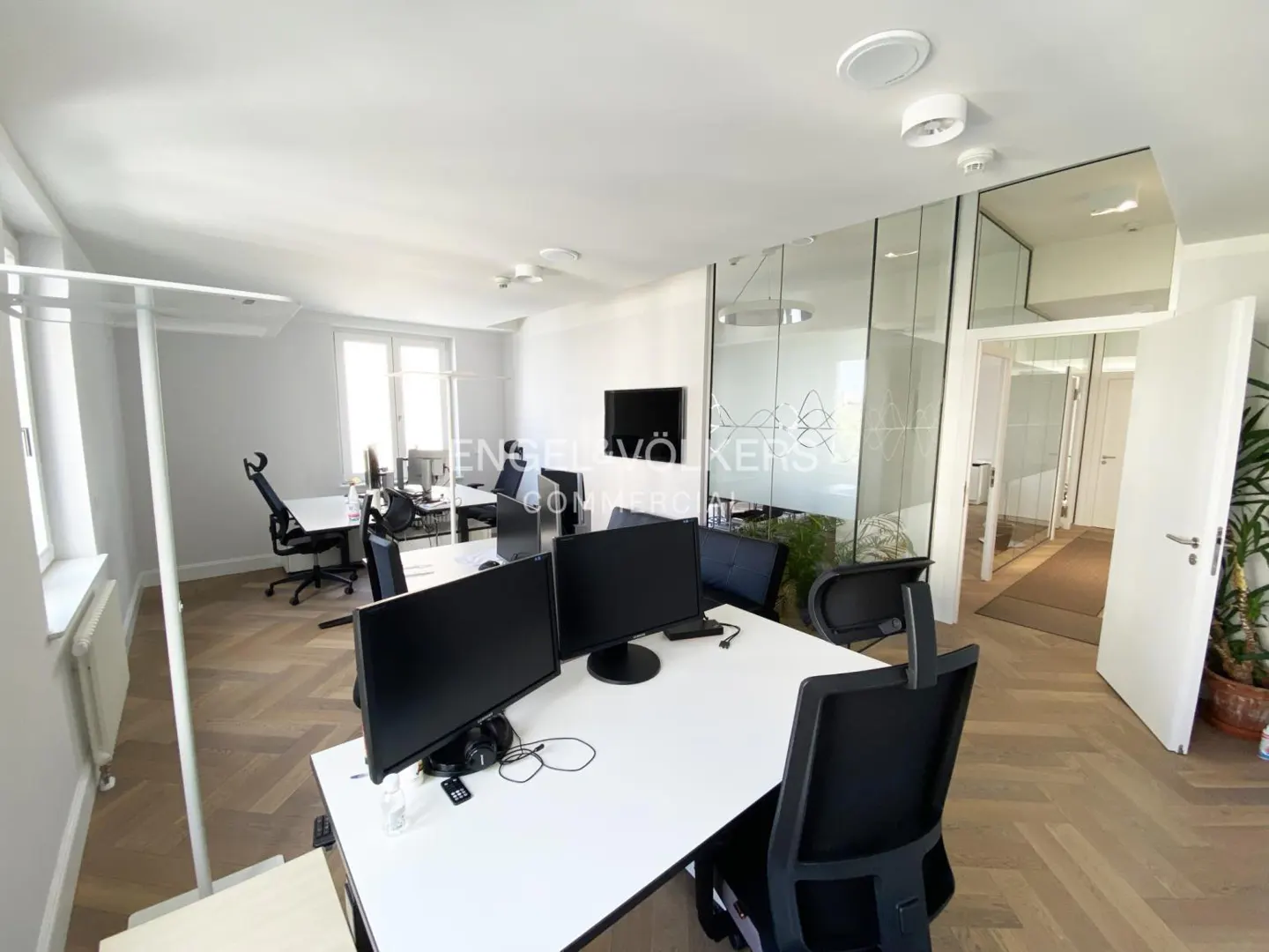 Bright office space with white walls, herringbone floors, and glass-walled conference room. Desks with monitors and black chairs are visible.