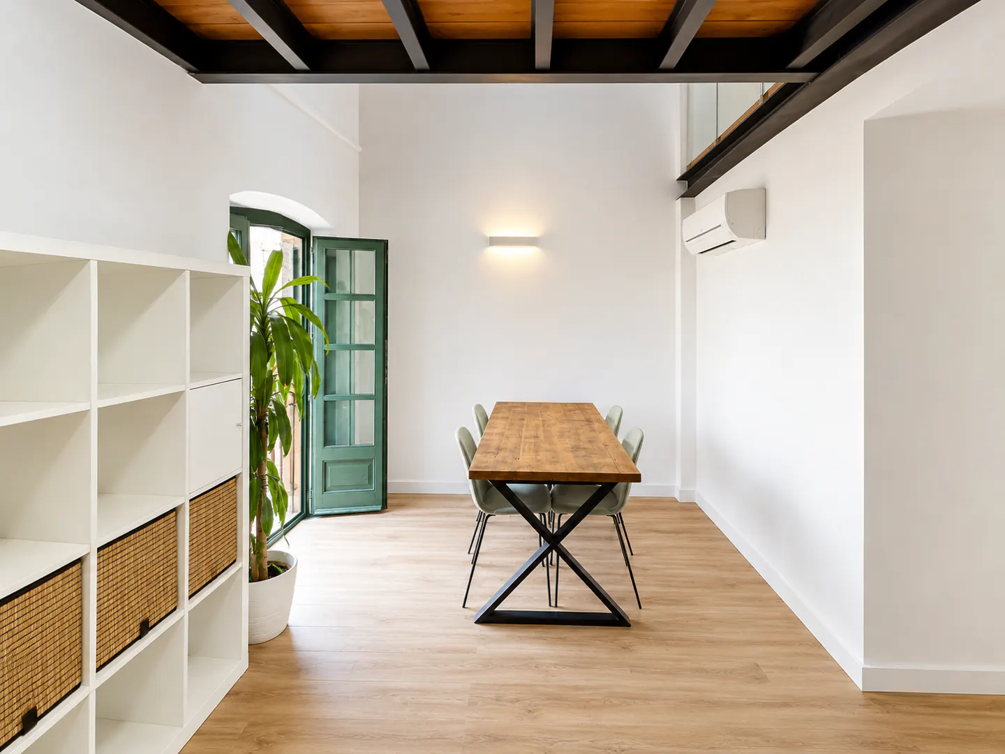 Bright, modern dining room with a wood table, green chairs, and a green door. A white shelf and potted plant are on the left.