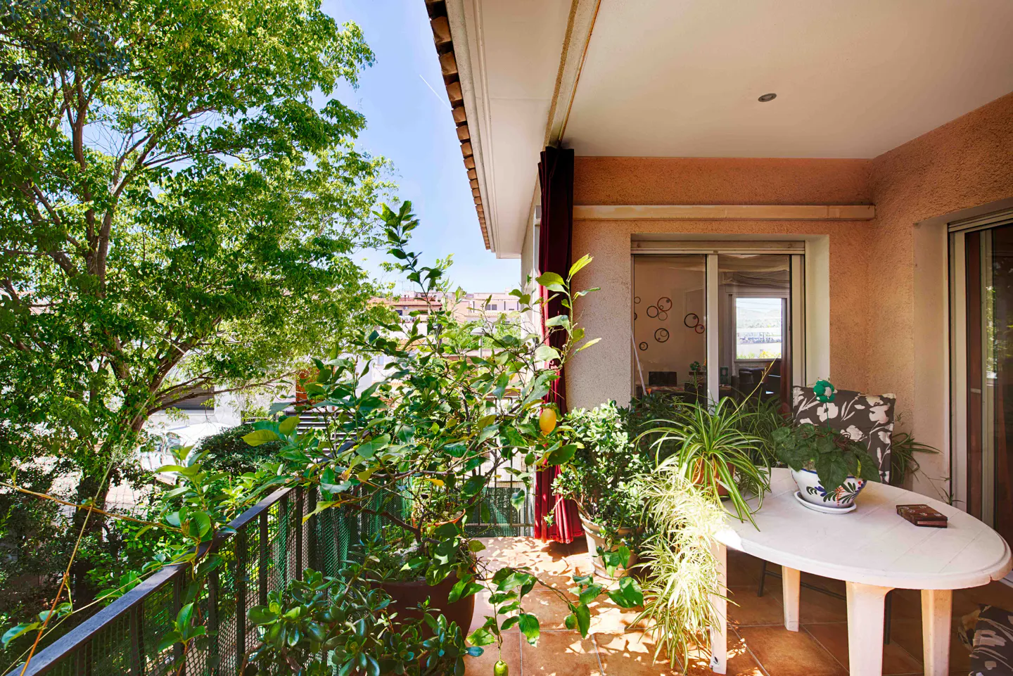 Balcony with plants, a small white table, and a view of trees and buildings in the distance. Red curtains are visible.