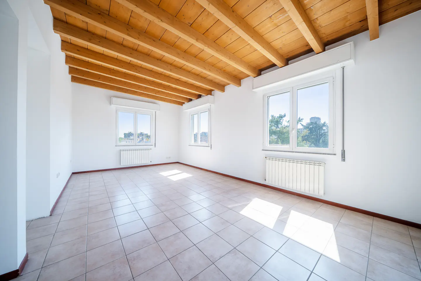 Bright, empty room with white walls, tile floor, and exposed wood beam ceiling. Three windows let in natural light.