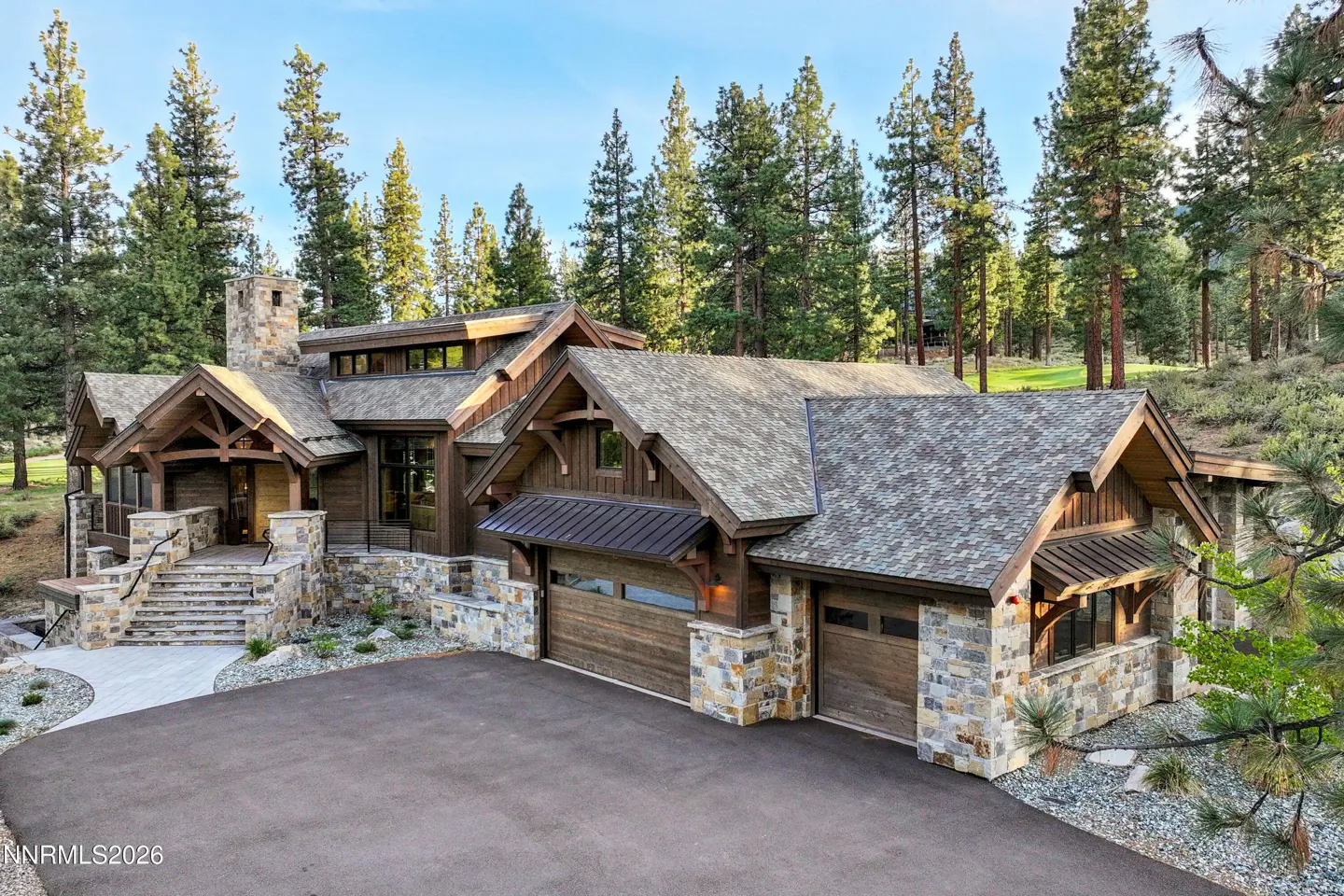 A large, brown, stone-accented house with a gray roof sits in front of tall green trees. A black asphalt driveway leads to the two-car garage.