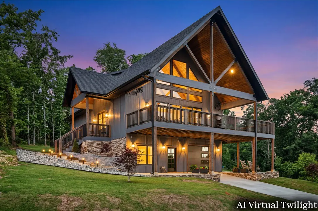 Modern A-frame home with dark siding, stone accents, and a large wooden deck at twilight, surrounded by trees.